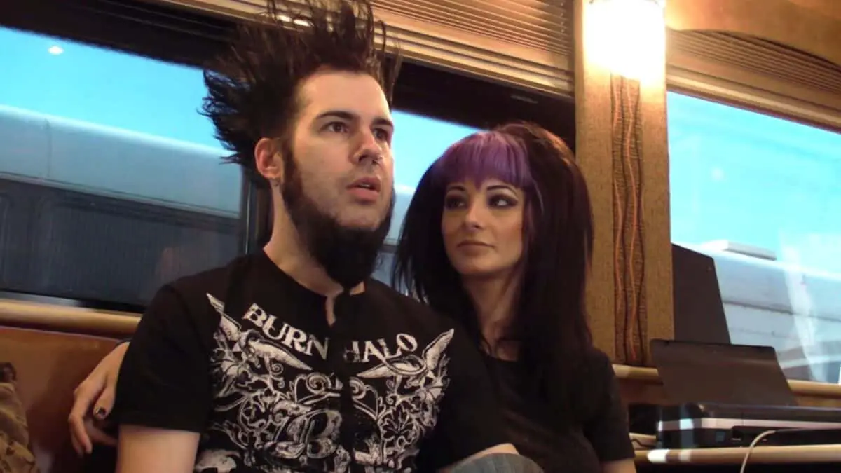 Spiky-haired man and woman with purple hair sitting together in a tour bus, enjoying a relaxed moment.